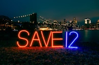 Brightly lit letters spelling 'SAVE12' appear in the foreground against an urban night skyline. The letters are illuminated with red and blue lights. A bridge adorned with glowing lights stretches across the background, leading to a cityscape filled with skyscrapers. The scene is reflected in a body of water, adding to the nighttime ambiance.