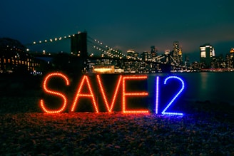 Brightly lit letters spelling 'SAVE12' appear in the foreground against an urban night skyline. The letters are illuminated with red and blue lights. A bridge adorned with glowing lights stretches across the background, leading to a cityscape filled with skyscrapers. The scene is reflected in a body of water, adding to the nighttime ambiance.