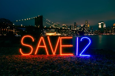 Brightly lit letters spelling 'SAVE12' appear in the foreground against an urban night skyline. The letters are illuminated with red and blue lights. A bridge adorned with glowing lights stretches across the background, leading to a cityscape filled with skyscrapers. The scene is reflected in a body of water, adding to the nighttime ambiance.
