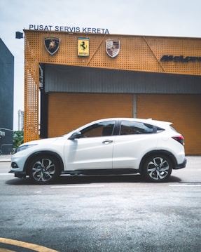 A white SUV is parked in front of a car service center with orange walls displaying luxury car brand logos including Lamborghini, Ferrari, and Porsche.