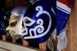 A collection of colorful lucha libre masks is displayed on wooden shelves. The masks feature intricate designs with metallic and vibrant colors, including blue, silver, and green. Next to the masks, there is a textile with blue, red, and white stripes.