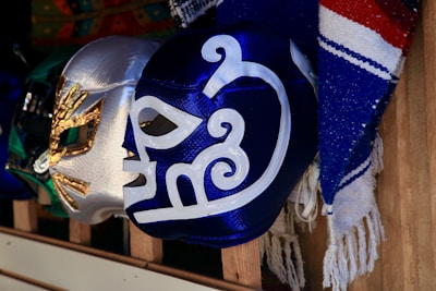 A collection of colorful lucha libre masks is displayed on wooden shelves. The masks feature intricate designs with metallic and vibrant colors, including blue, silver, and green. Next to the masks, there is a textile with blue, red, and white stripes.