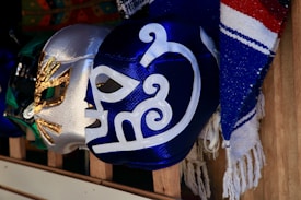 A collection of colorful lucha libre masks is displayed on wooden shelves. The masks feature intricate designs with metallic and vibrant colors, including blue, silver, and green. Next to the masks, there is a textile with blue, red, and white stripes.