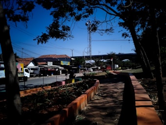 A public space featuring a paved pathway lined with orange borders and plants on either side. Tree branches with sparse foliage partially cover the scene. In the background, buildings with red-tiled roofs and a telecommunications tower are visible. A white truck is parked nearby under clear blue skies.