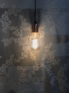A lit Edison-style light bulb hangs against a background of floral lace patterns, casting warm light that contrasts with the delicate flowers.