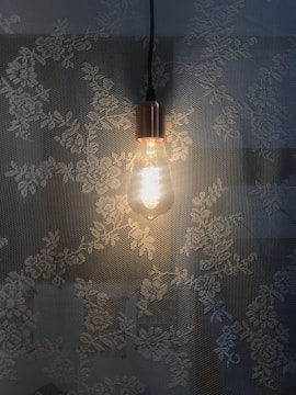A lit Edison-style light bulb hangs against a background of floral lace patterns, casting warm light that contrasts with the delicate flowers.