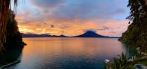 A picturesque lake at sunset with a serene expanse of water bordered by lush greenery. A prominent volcano is silhouetted against the vibrant sky, which transitions from warm oranges and pinks to cooler blues. The foliage on both sides adds depth to the tranquil scene.