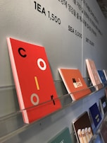 Colorful card designs are neatly displayed on a wall-mounted rack. Various cards feature different colors and text, with one prominently featuring the word 'color' in vibrant red with black and yellow accents. Above the display, there's text in Korean and numbers indicating pricing details.