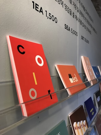 Colorful card designs are neatly displayed on a wall-mounted rack. Various cards feature different colors and text, with one prominently featuring the word 'color' in vibrant red with black and yellow accents. Above the display, there's text in Korean and numbers indicating pricing details.