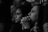 Two individuals appear focused and contemplative, sitting side by side in a dimly lit environment. Their hands are clasped together, resting near their faces, and their expressions suggest deep concentration or concern.