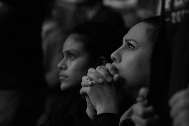 Two individuals appear focused and contemplative, sitting side by side in a dimly lit environment. Their hands are clasped together, resting near their faces, and their expressions suggest deep concentration or concern.