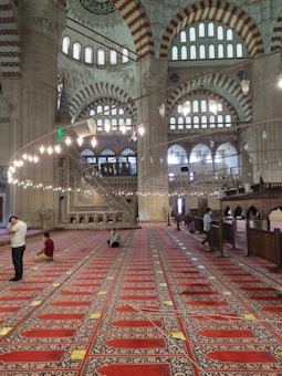 The image depicts a stunning interior of a mosque with high vaulted ceilings and intricate geometric designs. Light streams through decorated windows and chandeliers hang elegantly overhead. People are seen either seated or standing in prayer on richly patterned red and blue carpets stretching across the expansive hall.