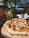 A freshly baked pizza topped with cheese and cooked meat sits on a wooden board. In the background, a glass of red cocktail is placed next to a green bottle adorned with string lights. The setting appears to be a cozy restaurant with brick walls and dim lighting.