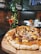 A freshly baked pizza topped with cheese and cooked meat sits on a wooden board. In the background, a glass of red cocktail is placed next to a green bottle adorned with string lights. The setting appears to be a cozy restaurant with brick walls and dim lighting.