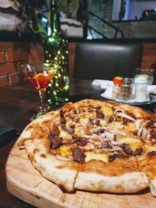 A freshly baked pizza topped with cheese and cooked meat sits on a wooden board. In the background, a glass of red cocktail is placed next to a green bottle adorned with string lights. The setting appears to be a cozy restaurant with brick walls and dim lighting.