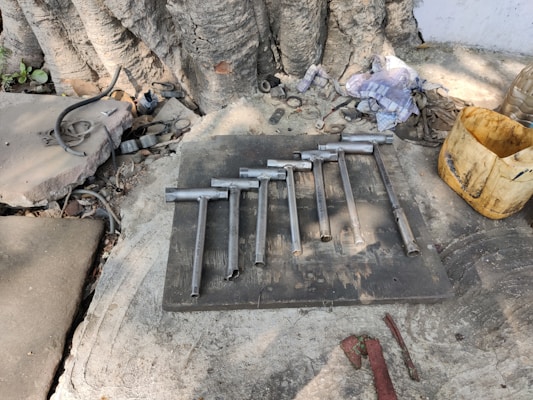 A set of metal tools resembling socket wrenches laid out on a flat surface. Nearby are worn cloths, a yellow container, and various mechanical parts scattered around. The scene is set outdoors against a large tree trunk with visible roots.