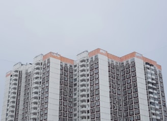 A high-rise apartment building with a modern architectural style is seen, featuring multiple balconies and windows arranged in a grid pattern. The upper part of the building has a distinctive orange trim, contrasting with the predominantly white and brown facade. The sky is overcast, giving the scene a muted ambiance.