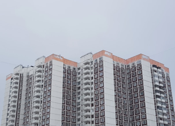 A high-rise apartment building with a modern architectural style is seen, featuring multiple balconies and windows arranged in a grid pattern. The upper part of the building has a distinctive orange trim, contrasting with the predominantly white and brown facade. The sky is overcast, giving the scene a muted ambiance.