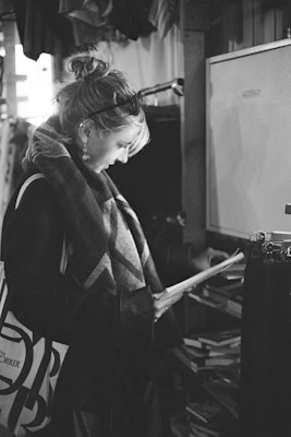 A black and white photograph captures a woman standing indoors, intently reading a document. She is dressed in a stylish coat and scarf, with her hair tied up. The environment suggests a shop or a library setting, with shelves of books visible in the background.