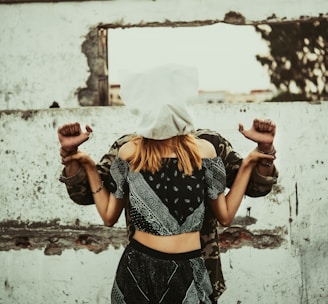A woman with a white cloth covering her head stands in front of a man whose hands are bound behind her back. They are positioned against a rough, textured wall with an opening, creating a sense of entrapment or confinement.