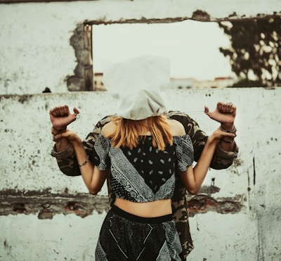 A woman with a white cloth covering her head stands in front of a man whose hands are bound behind her back. They are positioned against a rough, textured wall with an opening, creating a sense of entrapment or confinement.