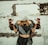 A woman with a white cloth covering her head stands in front of a man whose hands are bound behind her back. They are positioned against a rough, textured wall with an opening, creating a sense of entrapment or confinement.