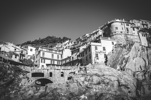 A black and white image depicting a coastal village with buildings perched on steep cliffs. The architecture reflects traditional Mediterranean style, with multiple stories and balconies. In the foreground, large rocks are visible, and a stone bridge connects part of the landscape. Some people can be seen exploring the rocky terrain.