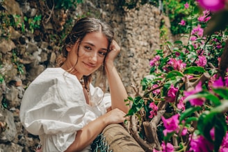A young woman in a white dress is leaning against a rustic wooden fence adorned with vibrant pink and purple flowers. She has a gentle smile on her face, with one hand resting on her cheek. The background features a stone wall, giving a rustic charm to the setting.