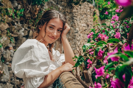 A young woman in a white dress is leaning against a rustic wooden fence adorned with vibrant pink and purple flowers. She has a gentle smile on her face, with one hand resting on her cheek. The background features a stone wall, giving a rustic charm to the setting.
