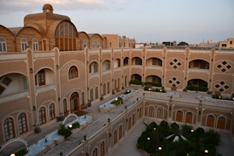 A multi-story building with an intricate design and arched windows, surrounded by a courtyard with greenery and pathways. The architecture features classic elements with a modern touch, and the building's facade is adorned with beige and brown tones. The courtyard has small garden areas with plants and shrubs.