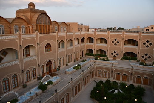 A multi-story building with an intricate design and arched windows, surrounded by a courtyard with greenery and pathways. The architecture features classic elements with a modern touch, and the building's facade is adorned with beige and brown tones. The courtyard has small garden areas with plants and shrubs.