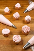Close-up of hands carefully piping pastel pink frosting onto a batch of freshly baked cupcakes.