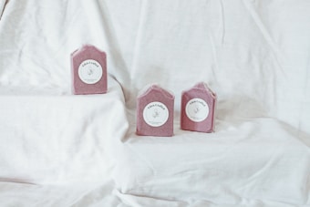 Three pink, handcrafted soap bars are placed on a soft white fabric backdrop. Each soap has a simple label with delicate illustrations and script, creating an elegant and artisanal presentation.