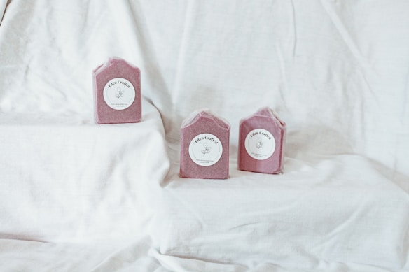 Three pink, handcrafted soap bars are placed on a soft white fabric backdrop. Each soap has a simple label with delicate illustrations and script, creating an elegant and artisanal presentation.