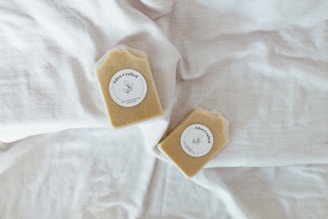Two bars of soap with labels reading 'Eden Crafted' are placed on a soft white fabric. The soap bars are a light beige color, and the fabric they rest on appears to be a bed sheet or similar textile.