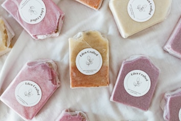 Several handmade soaps, each wrapped in paper and labeled 'Eden Crafted,' are arranged on a light fabric background. The soaps are in different colors, including shades of pink, beige, and brown. Each soap bar carries a round label with a simple botanical design and text indicating they are 100% handcrafted and locally made.