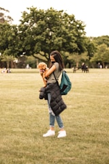 A person holding a small dog in a grassy park area, with trees in the background. The individual is wearing casual clothing, including jeans and sneakers, and has a green backpack. Others can be seen sitting and walking in the distance.