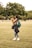 A person holding a small dog in a grassy park area, with trees in the background. The individual is wearing casual clothing, including jeans and sneakers, and has a green backpack. Others can be seen sitting and walking in the distance.