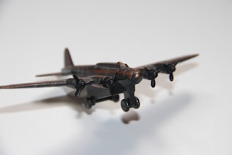 A close-up view of a vintage propeller airplane model with four propellers, casting a shadow on a light surface. The model is composed of a dark, possibly metallic material with low sheen.