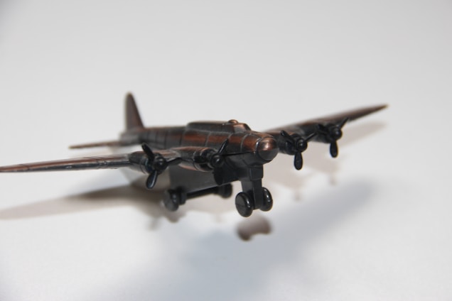 A close-up view of a vintage propeller airplane model with four propellers, casting a shadow on a light surface. The model is composed of a dark, possibly metallic material with low sheen.