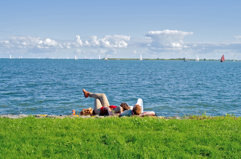 2 women lying on green grass field near body of water during daytime