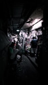 A narrow, dimly lit market alley filled with various goods. People are scattered throughout, some engaging in conversation or browsing items stacked on shelves and tables. The ceiling is low and lighting is sparse, creating an atmospheric and slightly mysterious ambiance.