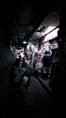 A narrow, dimly lit market alley filled with various goods. People are scattered throughout, some engaging in conversation or browsing items stacked on shelves and tables. The ceiling is low and lighting is sparse, creating an atmospheric and slightly mysterious ambiance.