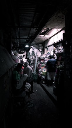 A narrow, dimly lit market alley filled with various goods. People are scattered throughout, some engaging in conversation or browsing items stacked on shelves and tables. The ceiling is low and lighting is sparse, creating an atmospheric and slightly mysterious ambiance.