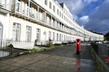 A row of elegant white terraced houses with intricate wrought-iron balconies lines a paved pathway. A bright red postbox stands prominently on the right beside the pavement, contrasting against the light-colored buildings. The sky is partly cloudy, casting a mix of sunlight and shadow on the scene.
