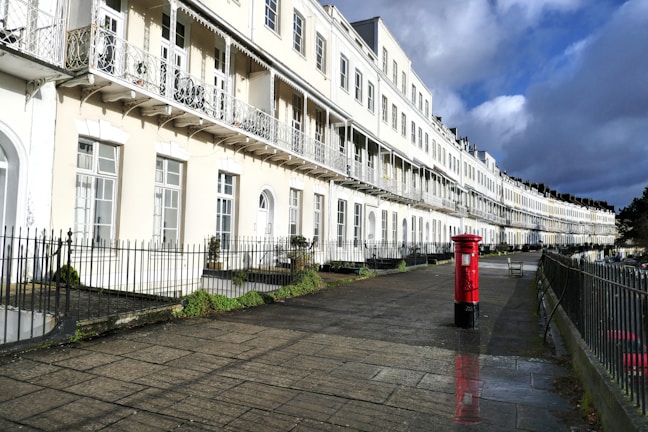 A row of elegant white terraced houses with intricate wrought-iron balconies lines a paved pathway. A bright red postbox stands prominently on the right beside the pavement, contrasting against the light-colored buildings. The sky is partly cloudy, casting a mix of sunlight and shadow on the scene.
