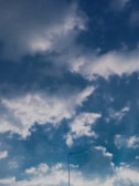 A partly cloudy sky with a mix of white and gray clouds spread across a blue background. In the lower part of the image, a long, thin antenna is visible, extending upwards.