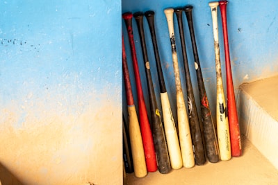 Several baseball bats are arranged upright against a textured blue wall. The bats have a mix of black, white, and red colors. The wall shows a gradient of blue and beige, creating a visually appealing backdrop. There is a step to the right.