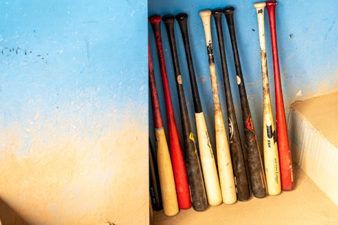 Several baseball bats are arranged upright against a textured blue wall. The bats have a mix of black, white, and red colors. The wall shows a gradient of blue and beige, creating a visually appealing backdrop. There is a step to the right.