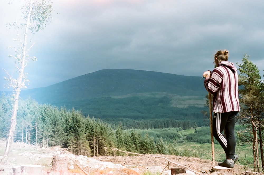 man in brown jacket standing on rock near green trees during daytime